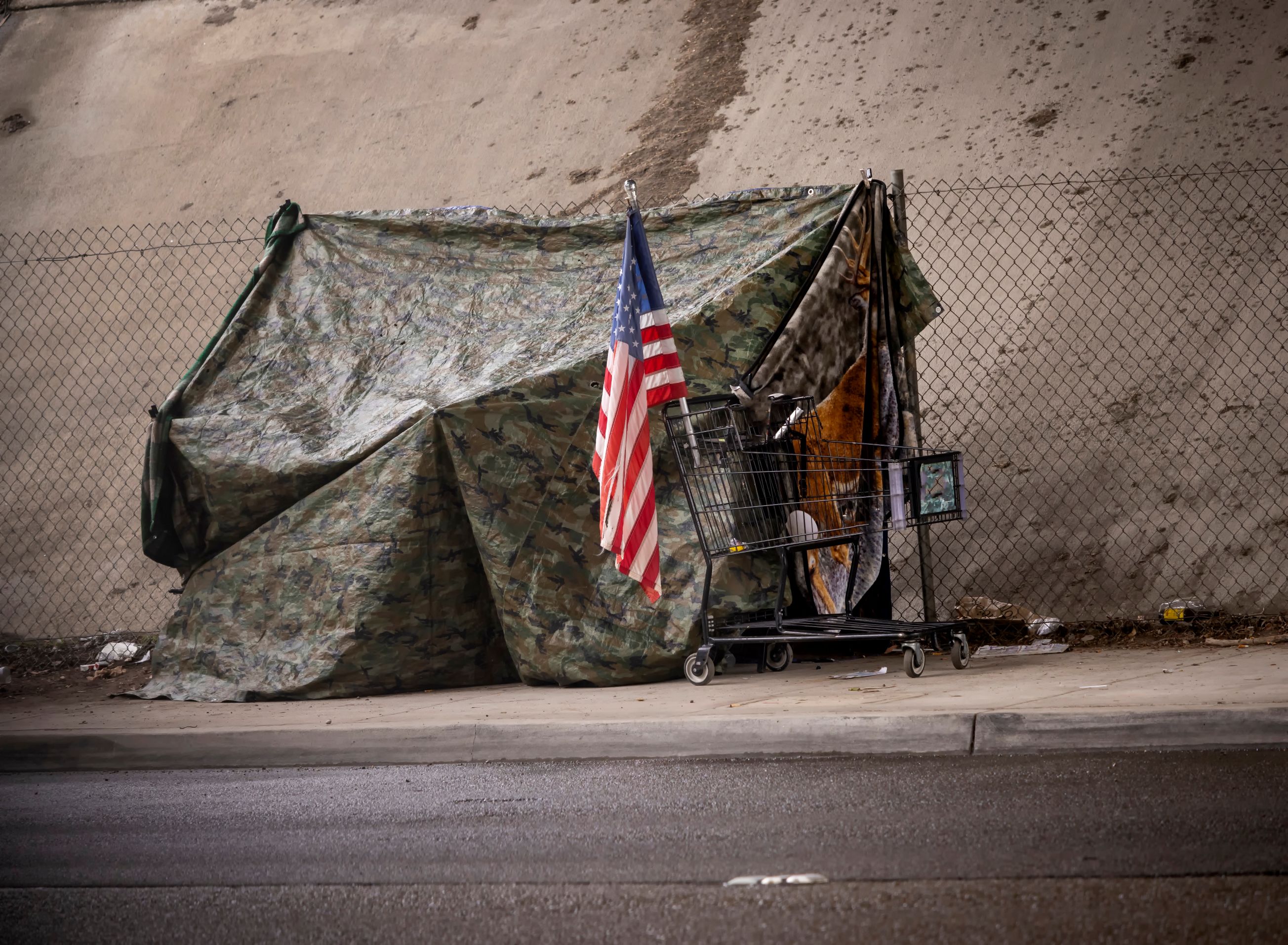 An American flag at a homeless tent made of camouflage tarp at a road underpass.